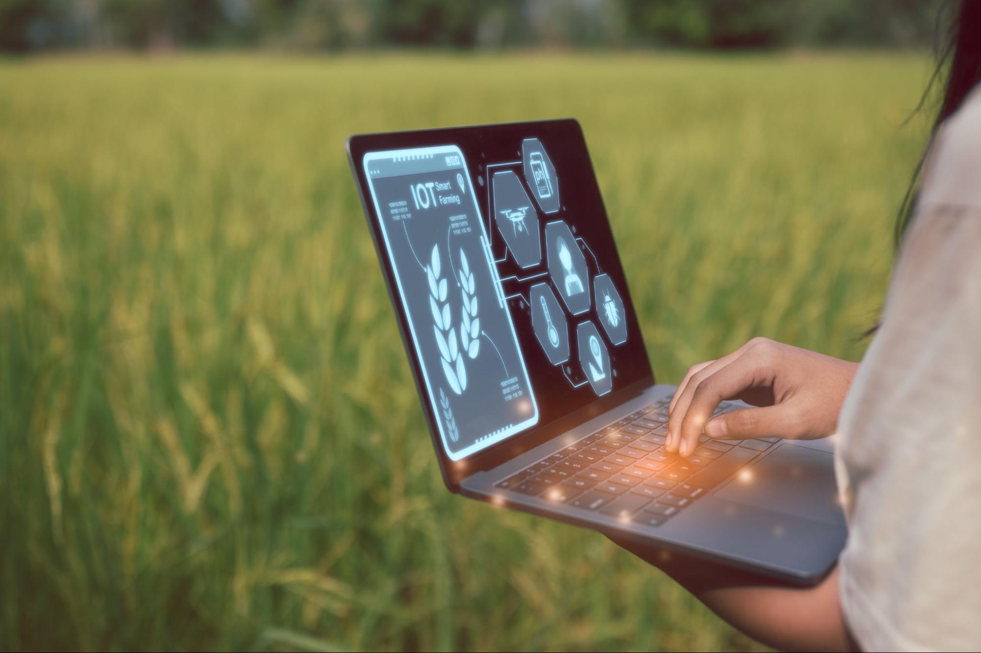 A person using a laptop in a green field, displaying smart farming analytics with icons for IoT, crops, and sensors